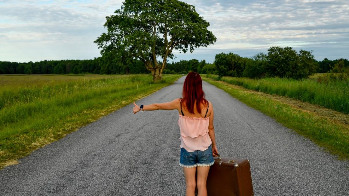 A woman with red hair hitchhiking on a country road in Estonia with a vintage suitcase.