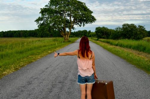 A woman with red hair hitchhiking on a country road in Estonia with a vintage suitcase.