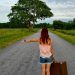 A woman with red hair hitchhiking on a country road in Estonia with a vintage suitcase.